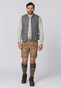 Grau-linener Weste über einem hellgrünen Hemd, kombiniert mit braunen Lederhosen mit floralen Stickereien, grauen gestrickten Kniestrümpfen und braunen Stiefeln.