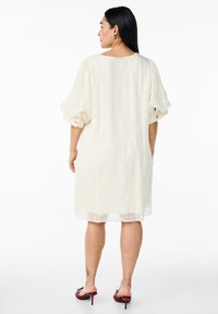 Robe courte crème en tissu texturé, avec des manches courtes volumineuses et des détails de boutons. Portée avec des sandales à talons rouges.