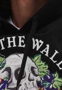 Sweat à capuche noir avec un grand motif de crâne brodé entouré d'accents floraux colorés. Le texte indique "THE WALL." Comprend un cordon de serrage.