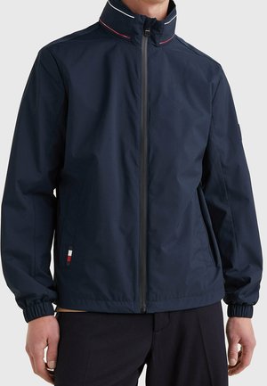 Mann trägt eine marineblaue Jacke mit Reißverschluss, hohem Kragen, elastischen Bündchen und einem kleinen rot-weiß-blauen Etikett nahe der linken Tasche.