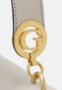 Fibbia circolare dorata con il logo 'G' in rilievo attaccata a una cinghia liscia e grigia chiara. Anello di metallo collega la cinghia a una superficie bianca.