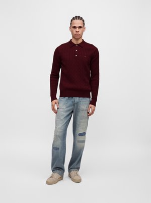 Ung mand med fletninger, iført en mørkerød striktrøje, lyseblå patched jeans og beige sneakers, står mod en ensfarvet baggrund.