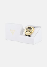 Reloj dorado con esfera negra y correa a juego, que cuenta con tres subesferas, alojado en una caja blanca con un logo dorado.
