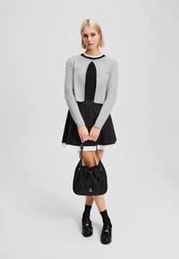 Cardigan grigio corto con bordo nero, abbinato a un vestito nero e a una borsa a secchiello nera. Presenta un colletto rotondo e chiusura a bottoni.