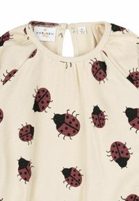 Top in tessuto bianco sporco con motivi di coccinelle rosse e nere, dotato di un'apertura a chiave sul retro e dettagli arricciati sulle spalle.