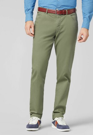 Homme portant un pantalon vert olive, une chemise bleue rentrée, une ceinture en cuir marron et des baskets bleues, blanches et marron, debout devant un fond uni.