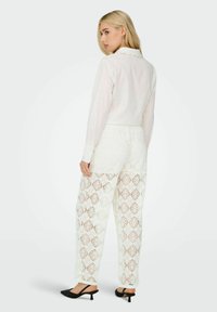 Blusa bianca con dettagli in perla, abbinata a pantaloni bianchi larghi in pizzo con motivo a diamante. L'outfit è completato da mules nere con tacco.