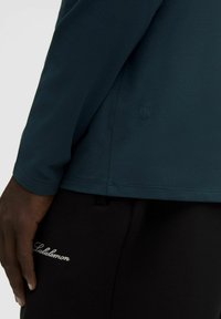 Dlhý rukáv tyrkysovej košele s zaobleným spodným okrajom, spárovaný s čiernymi nohavicami s bielym kurzívnym logom "Lululemon" a štruktúrovanou tkaninou.