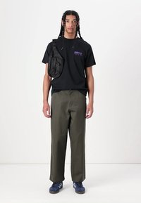 Schwarzes T-Shirt mit lila grafischem Text, olivgrüneweite Hosen und blaue Sneaker, begleitet von einer schwarzen Umhängetasche.