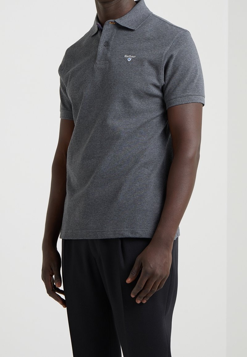 Homme portant un polo à manches courtes gris foncé avec un petit logo Barbour sur la poitrine et un pantalon noir, debout devant un fond uni.