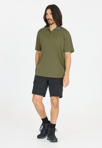 Olivgrön polo med krage, tillsammans med svarta shorts med dragkedjefickor och svarta vandringskängor.