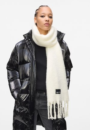 Personne portant un manteau matelassé noir brillant, un pull noir, un jean foncé et une épaisse écharpe frangée blanche enroulée autour du cou.
