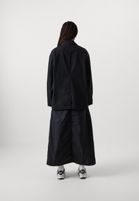 Svart oversized jakke med knepping, kombinert med et langt svart skjørt. Modellen har på seg mørke joggesko med et enkelt, moderne design.