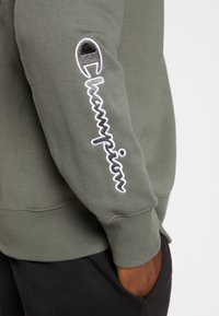 Manica di una felpa verde oliva con il logo "Champion" ricamato in bianco e nero, mano infilata nella tasca scura dei pantaloni.