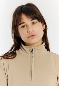 Bežový zipsový top s vysokým golierom, s čiernym zipsom a jemne embosovaným logom na prednej strane. Hladká textúra látky.