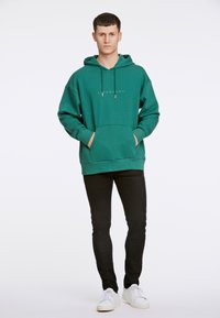 Grön hette med framficka, dragsko på huvan och silverlogotyp. Bärs med svarta smala jeans och vita sneakers. Enkel och avslappnad design.