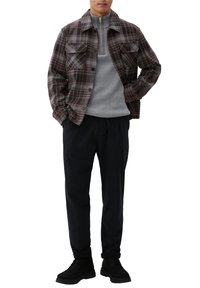 Homme portant une veste boutonnée à carreaux marron sur un pull à fermeture éclair gris, pantalon noir et chaussures noires, debout les mains dans les poches.