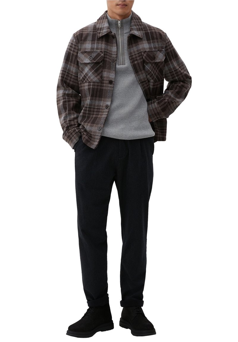 Homme portant une veste boutonnée à carreaux marron sur un pull à fermeture éclair gris, pantalon noir et chaussures noires, debout les mains dans les poches.