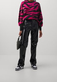 Persona che indossa pantaloni neri lucidi, sneakers bianco e nere, un pullover a righe rosa e nero, e tiene in mano una piccola borsa nera.