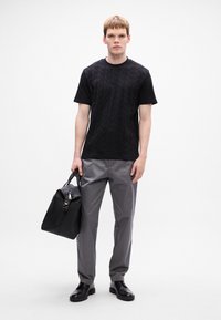 Czarny t-shirt w wzory, szare spodnie i czarne skórzane buty. Model trzyma czarną torbę z zamkiem błyskawicznym i paskami.
