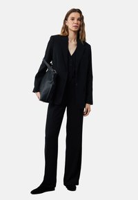Zwarte op maat gemaakte blazer over een bijpassend driedelig pak, met wijde broekspijpen en een gebuttonde vest, gecombineerd met een zwarte schoudertas.