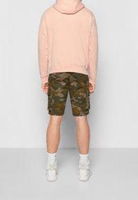 Ljusrosa huvtröja med ribbad nederkant, kombinerad med gröna camouflagecargo-shorts med flera fickor, b ärs med vita sneakers.