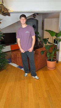 Mladi moški v vijolični majici in črnih hlačah smehlja se v zaprtih prostorih ob okrašeni božični jelki, veliki rastlini in rjavi kavču.