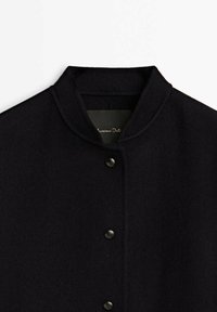 Black wool blend jacket features a stand collar, front button closure, and a textured finish. Simple and sleek design without patterns.