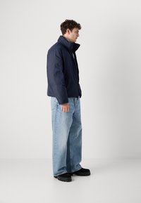 Marine blauwe pufferjack met hoge kraag, gecombineerd met lichtblauwe wijde jeans en zwarte chunky schoenen, staande aan de linkerkant tegen een effen achtergrond.