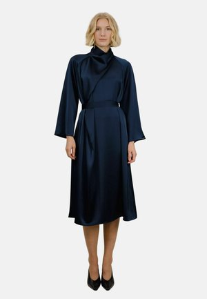 Mørkeblå sateng midi-kjole med drapert utringning og lange ermer, innsnevret midje, og skjørt som går under kneet. Svarte hæler.