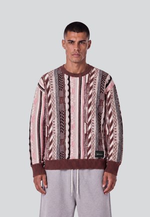 Mann trägt einen gemusterten Strickpullover mit braunen, rosa und cremefarbenen vertikalen Streifen und graue Kordelzughose vor einem schlichten Hintergrund.