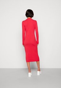 Femme aux cheveux courts et foncés portant une robe midi rouge à manches longues ajustée et des chaussures à talons blancs, se tenant devant un fond uni.