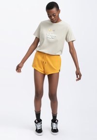 Lichtgrijze T-shirt met een kleurrafie, gecombineerd met felgele shorts. Zwarte hoge sneakers met witte veters en groene sokken.