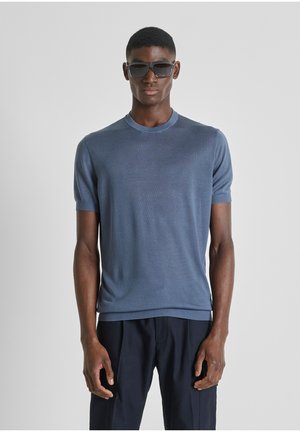 Uomo con occhiali da sole scuri, maglietta a maniche corte blu in maglia e pantaloni scuri, in piedi davanti a uno sfondo grigio uniforme.