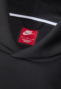 Černá mikina Nike s kapucí, vybavená bílým zipem, měkkou texturou látky a červenou štítkem zobrazujícím informace o velikosti a výrobě.