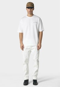 Hvid bomuld t-shirt med korte ærmer og logo foran, parret med hvide denim jeans og sorte ankelstøvler, sat mod en ensfarvet baggrund.