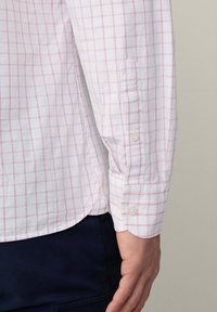 Camicia bianca con motivo a griglia rosa, maniche lunghe con polsini a bottone, realizzata in tessuto leggero, che mostra i dettagli delle cuciture laterali e dell'orlo.