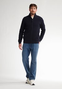 Marineblauwe geribbelde zip-up trui met hoge kraag, gedragen over een crèmekleurig shirt, gecombineerd met blauwe spijkerbroeken en witte sneakers.