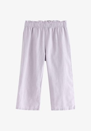 Smilende barn med krøllet hår iført hvid øjelygert top uden ærmer, lys lilla bukser med vide ben og hvide sko, stående på ensfarvet baggrund.