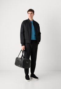 Zwarte bomberjack over een teal shirt, zwarte broek en zwarte sneakers. Houdt een zwarte sporttas vast met zilveren hardware en logo.