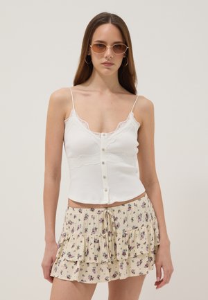 Mujer con gafas de sol redondas, pendientes de aro, una camiseta de tirantes blanca con adornos de encaje y botones, y una minifalda floral de capas, de pie frente a un fondo liso.