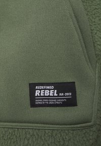 Zielony materiał z teksturowaną wstawką z polaru; czarna metka z napisem "REDEFINED REBEL RR-2013" w białym kolorze.
