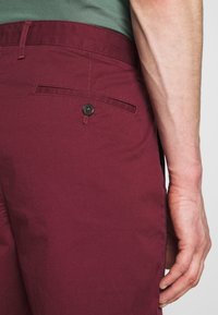 Pantaloni corti bordeaux con una texture liscia, dotati di una tasca posteriore e un dettaglio con bottone nero, mostrati in primo piano accanto a una mano.
