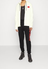 Sudadera amarilla claro con cremallera y acentos negros, combinada con pantalones de chándal negros que presentan la impresión en rojo de 'HUGO'. Botas negras completan el look.