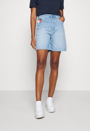 Persoon draagt lichtblauwe spijkershorts met knopen, een navy top en witte sneakers, staand tegen een effen lichte achtergrond.