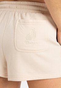 Beige shorts feature a soft texture, gathered elastic waistband, and a back pocket with subtle cactus embroidery and logo detail.