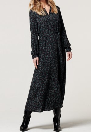 Žena v dlouhých černých šatech s zeleným květinovým vzorem, s opaskem v pase a černými kozačkami nad kolena, stojící před jednobarevným pozadím.