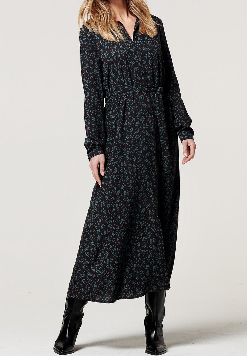 Femme portant une robe noire à manches longues avec un imprimé floral vert, une ceinture à la taille, et des bottes noires montant jusqu'aux genoux, debout devant un fond uni.