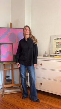 Camisa preta de botão com um corte descontraído, combinada com calças jeans azuis largas e sapatos pretos. O fundo apresenta uma pintura abstrata em tons de rosa.