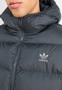 Fekete puffernadrág magas gallérral, cipzáras záródással és hímzett adidas logóval a mellkasán. Simább, könnyű anyagból készült.